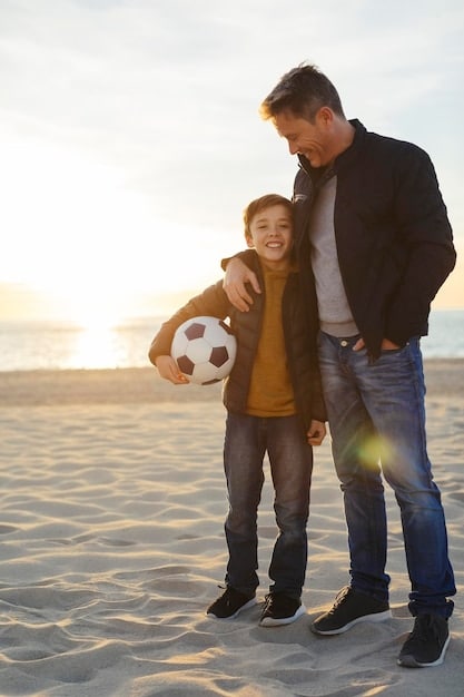 A father and his son, both smiling, standing side-by-side after a soccer practice. The boy is holding a soccer ball and looks proud, while the father is giving him a supportive pat on the back. The background shows other children and parents, creating a sense of community and positive involvement in youth sports. The focus is on the bond between father and son and the positive reinforcement of effort.
